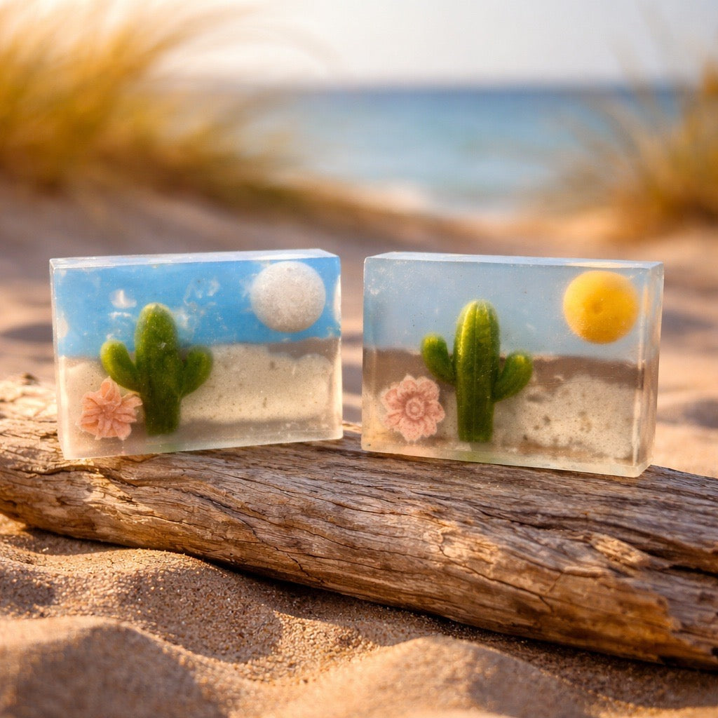 Two small square soaps sculptures with desert scenes on a sandy surface with blurred beach background